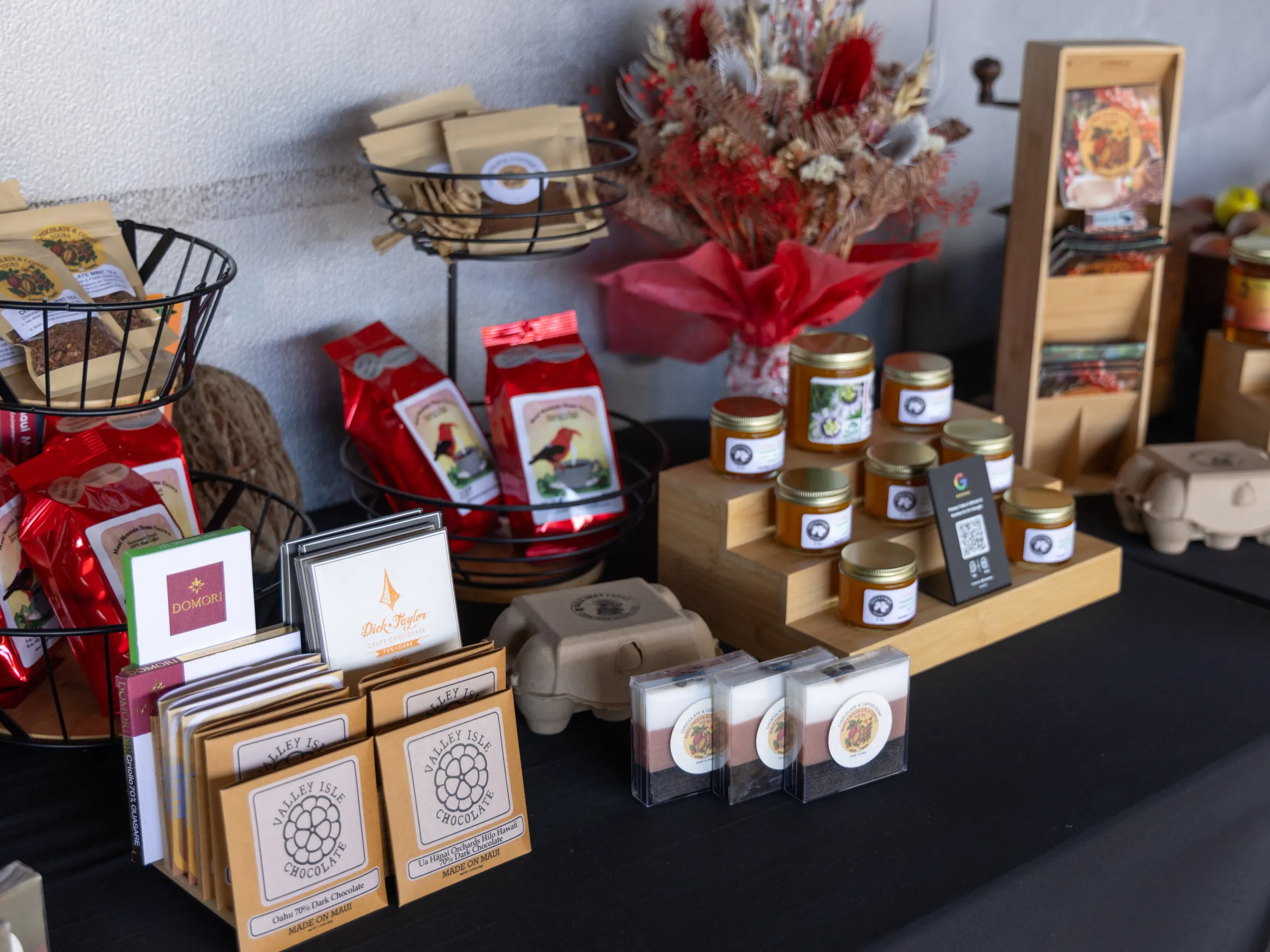 Display of assorted packaged foods and jars on a black tablecloth with a floral arrangement.