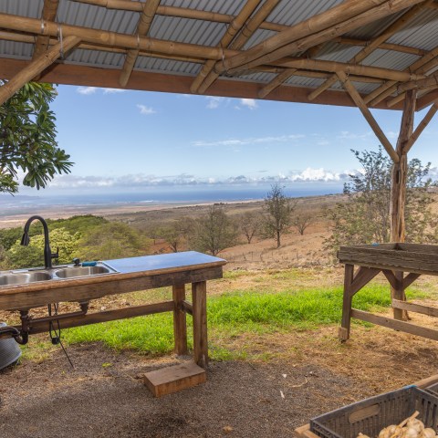 Outdoor kitchen setup under a wooden canopy with a scenic view of hills and ocean.