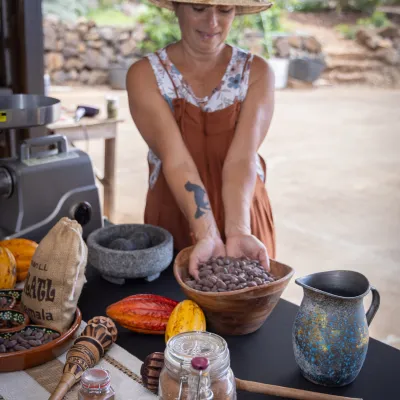 Woman in straw hat holding cocoa beans, with cacao pods and utensils on a table.
