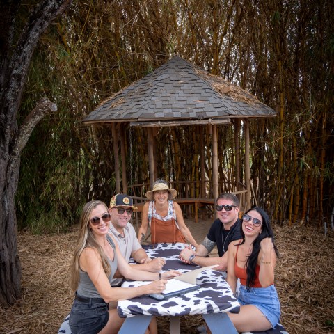 Five people sit at a picnic table near a wooden gazebo surrounded by bamboo.