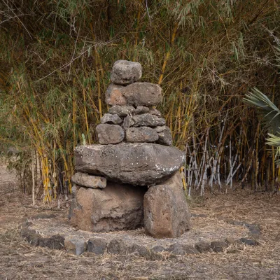 Stone cairn surrounded by bamboo and palm leaves, with parked cars in the background.