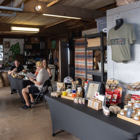 Inside market with products on display tables and people seated, concrete floor and wooden ceiling.