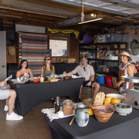 People seated at tables with food items in a workshop, with a woman speaking to the group.