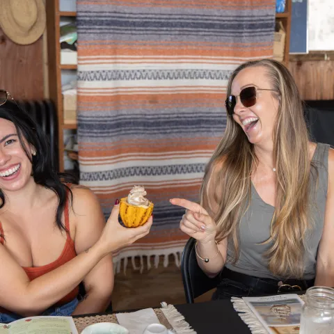 Two women laughing, one holding a decorated squash, in a cozy room.