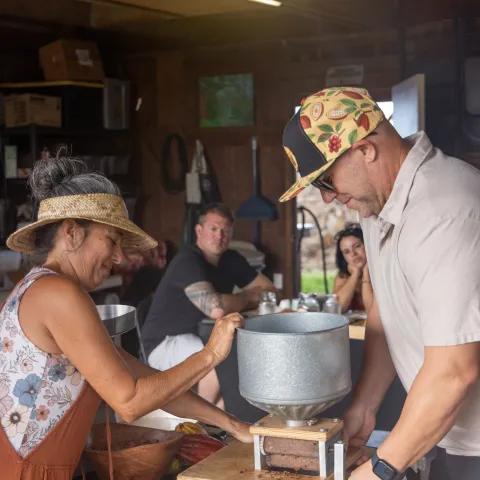 Two people operate a manual grinder in a workshop, with onlookers seated at a table.