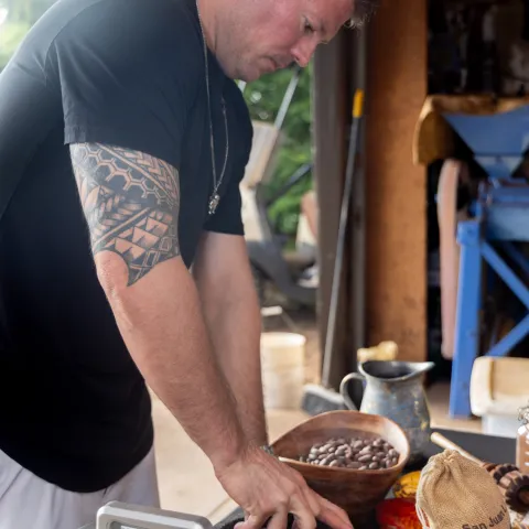 Man grinding cocoa beans with a mortar and pestle on a table with cacao pod and grinder.