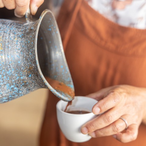 Person pouring chocolate into a white cup from a rustic metal pitcher.