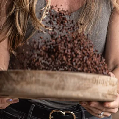 Person sifting cocoa beans in a wooden tray, wearing a sleeveless shirt.
